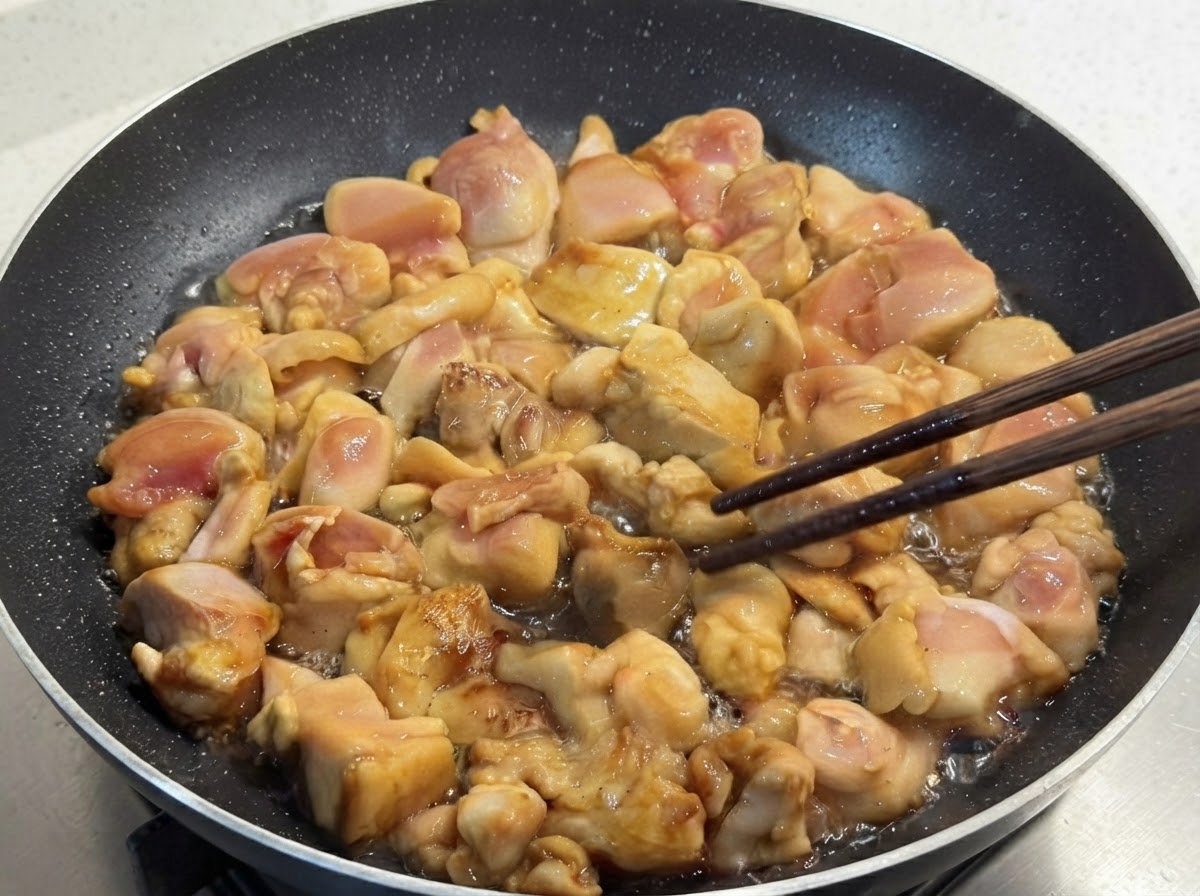 Trozos de pollo marinados friéndose en una sartén y siendo volteados cuidadosamente usando palillos de madera.