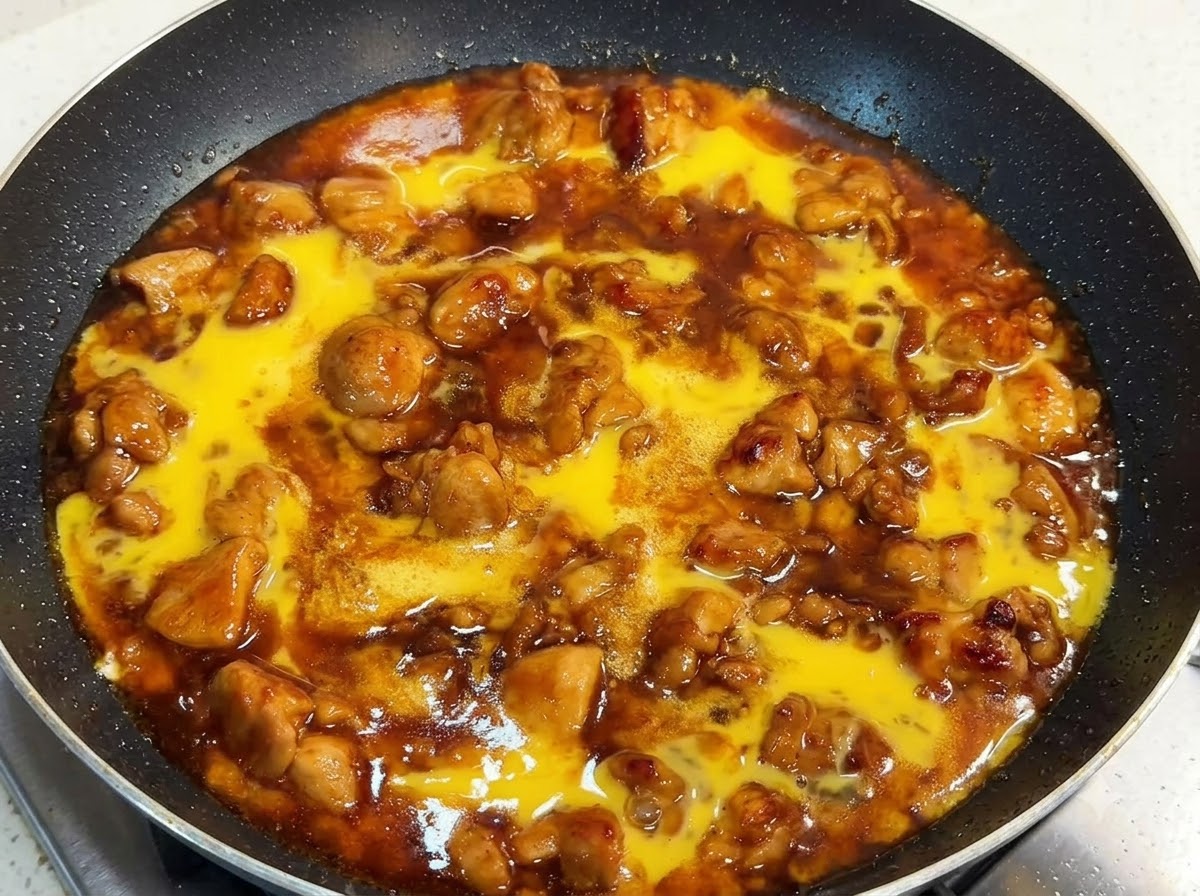 Una sartén llena de trozos de pollo y salsa rica cubierta con líquido de huevo batido crudo, tapada con una tapa.