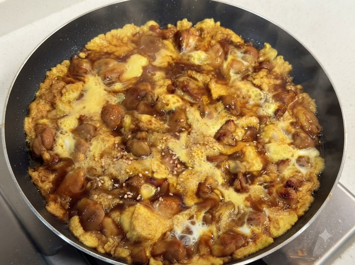 Una sartén caliente llena de pollo tierno cocinado y huevo sedoso en una salsa oscura sabrosa, adornada con semillas de sésamo blanco.