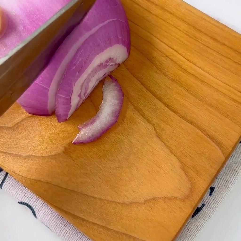 Una cebolla morada siendo cortada cuidadosamente en tiras finas sobre una tabla de cortar de madera con un cuchillo.