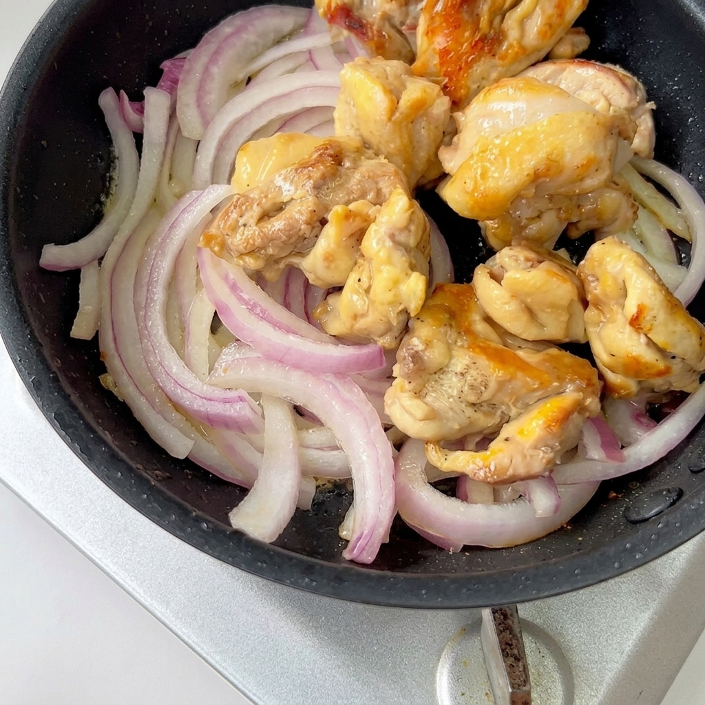 Trozos de pollo cocidos y cebollas moradas cortadas cociéndose a fuego lento en una sartén con salsa.
