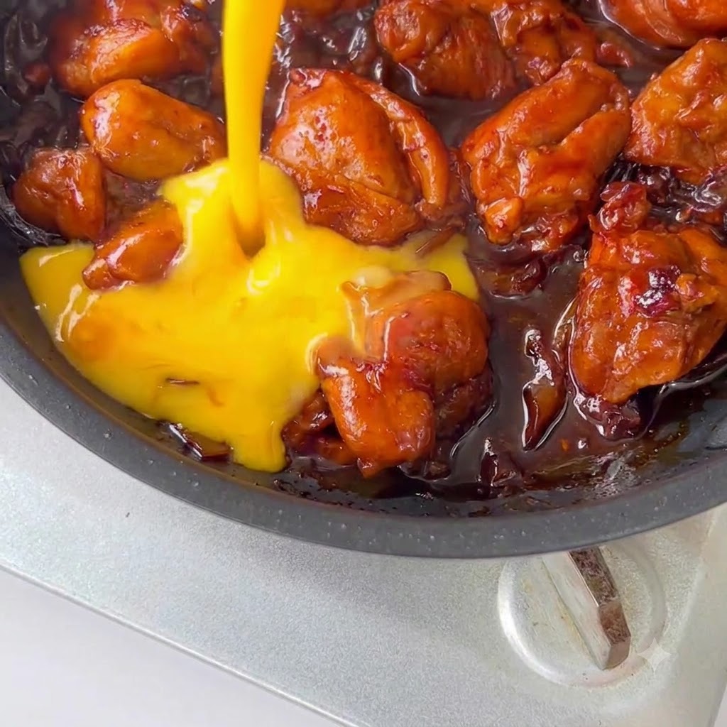 Huevos batidos siendo vertidos sobre el pollo y la salsa en una sartén.