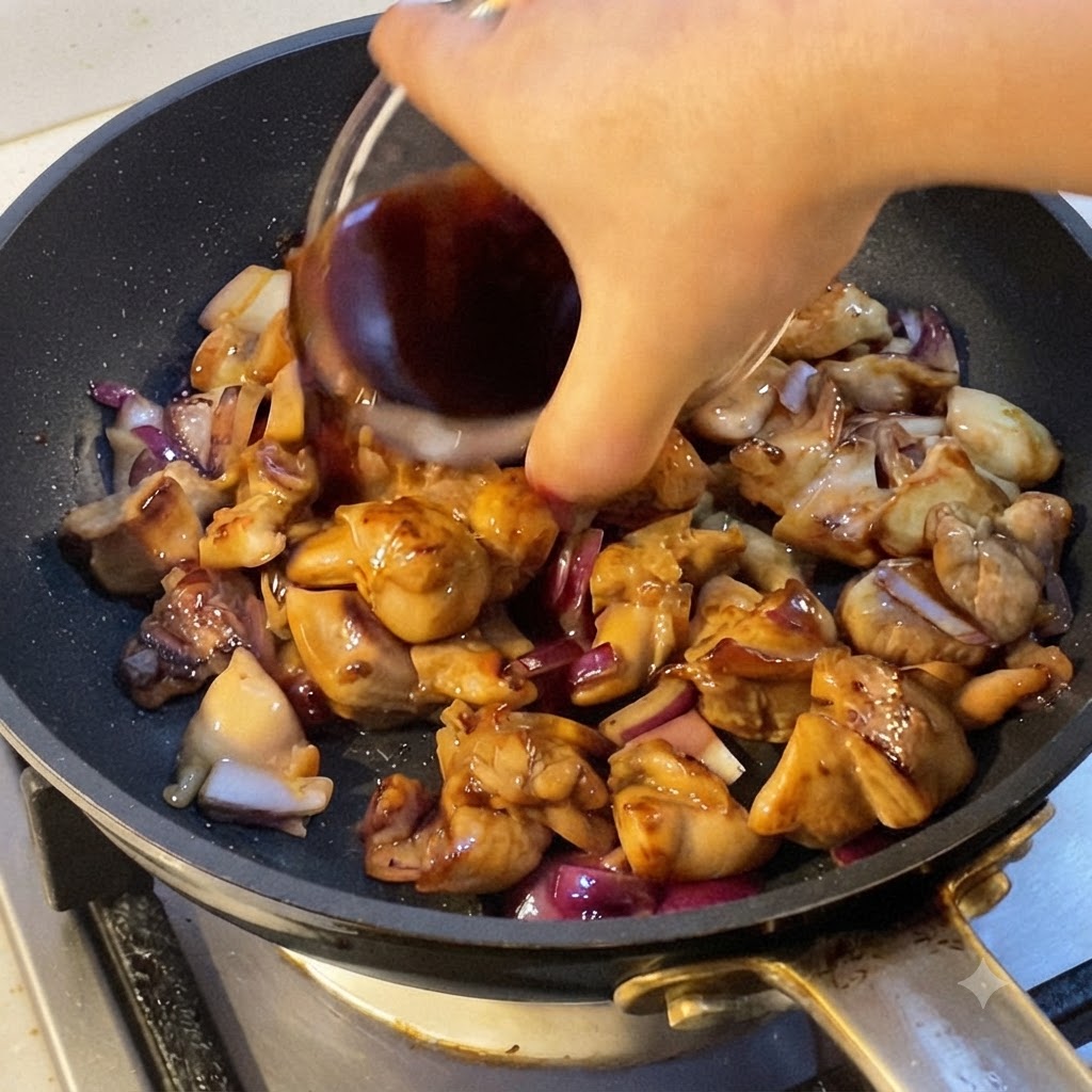 Una mano vertiendo salsa de cocina oscura en una sartén llena de trozos de pollo cocinado y trozos de cebolla roja.