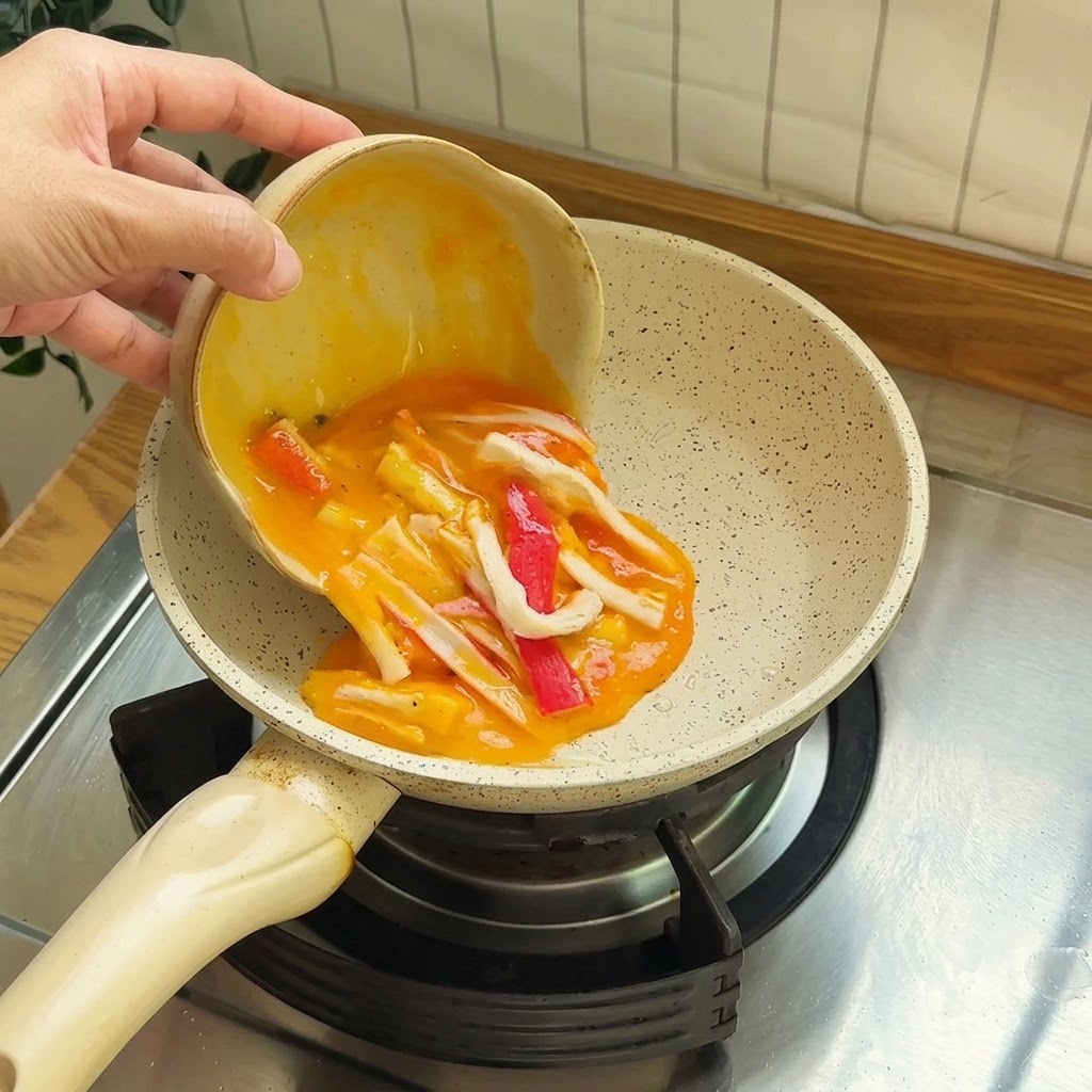 Huevos batidos mezclados con trozos rojos y blancos de palitos de cangrejo siendo vertidos en una pequeña sartén antiadherente.