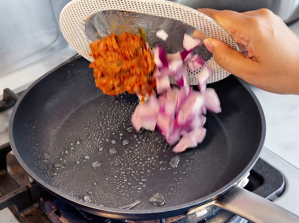 Cebollas moradas picadas y kimchi rojo siendo vertidos desde un plato colador en una sartén caliente con gotas de aceite.