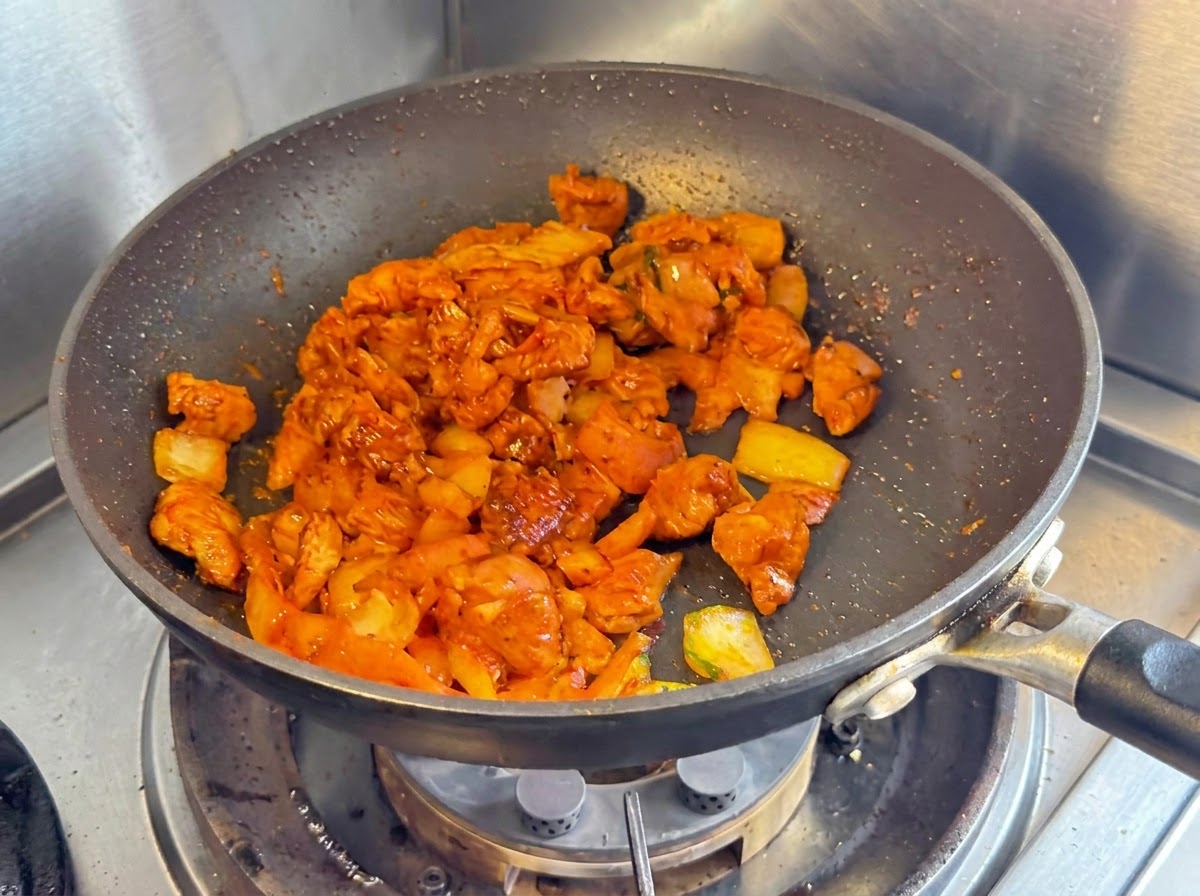 Trozos de pechuga de pollo, cebollas y kimchi picado salteándose juntos en una sartén antiadherente oscura.