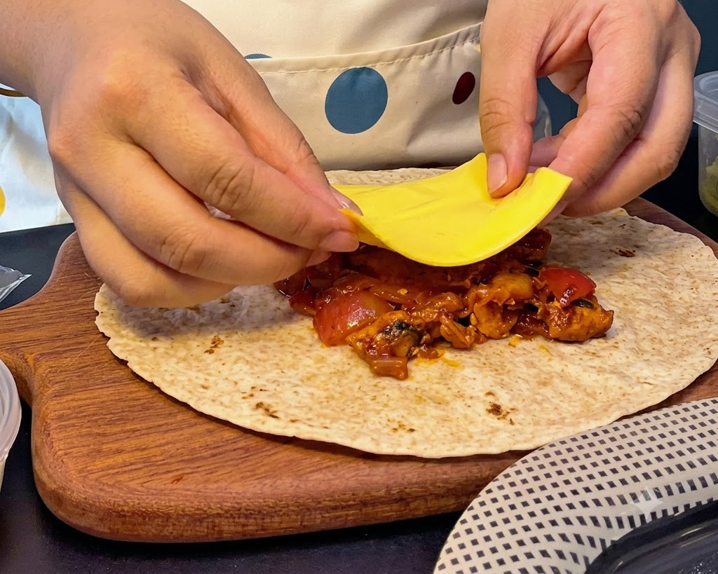 Manos colocando una rebanada cuadrada de queso amarillo sobre una pila de relleno caliente de pollo y kimchi en un pan plano.
