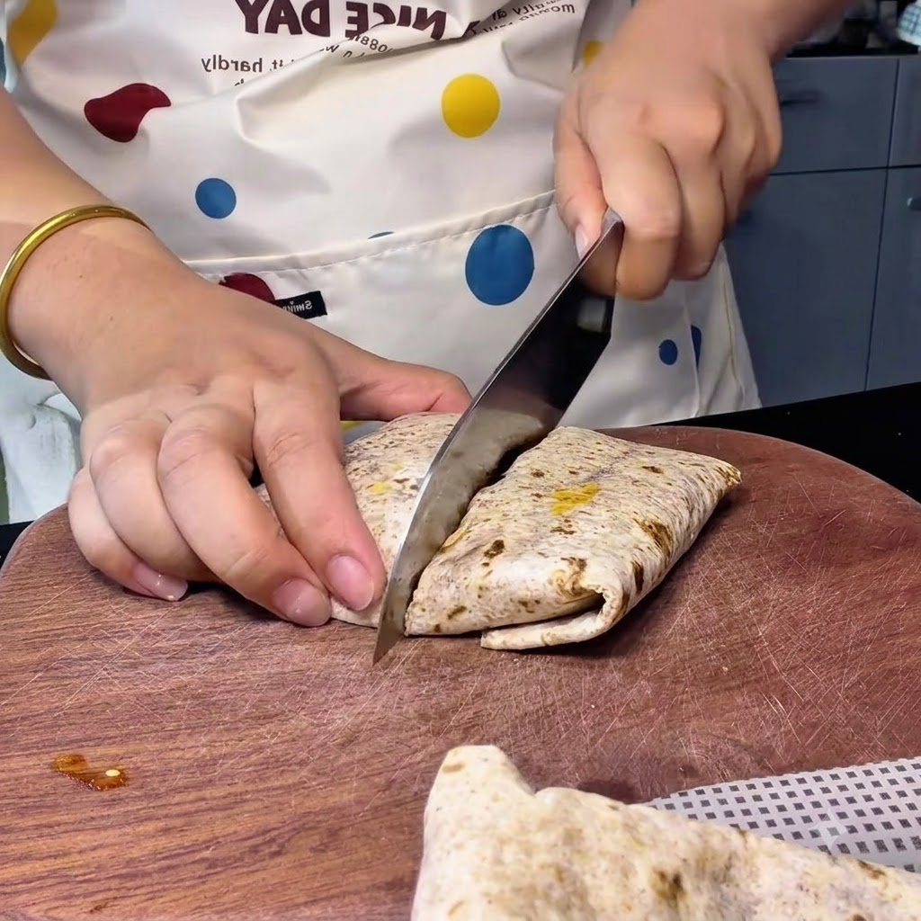 Una persona usando un cuchillo de chef grande para cortar un wrap integral dispuesto diagonalmente en una tabla de cortar de madera.