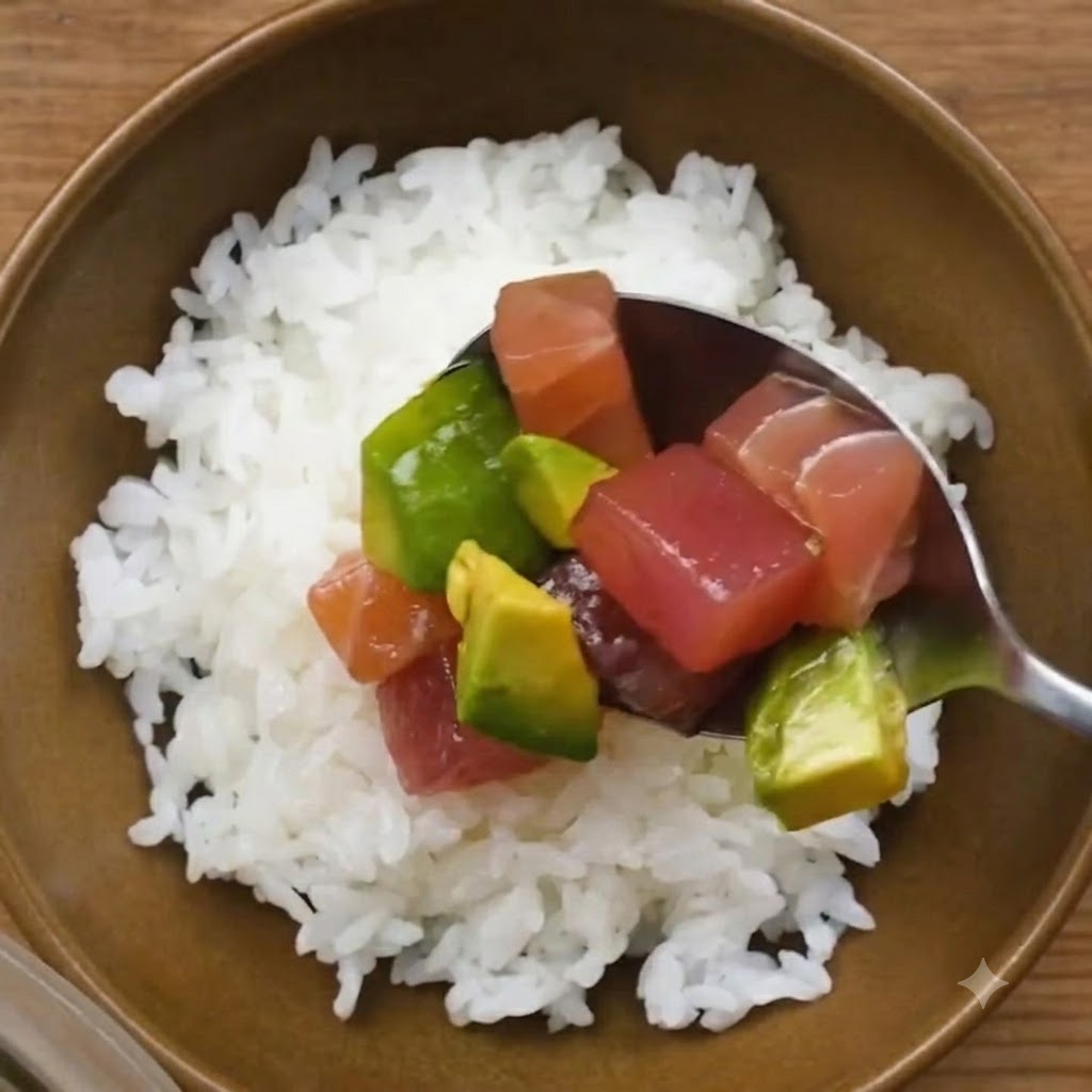 Una cucharada de atún marinado y aguacate siendo colocada sobre un tazón de arroz blanco para sushi.
