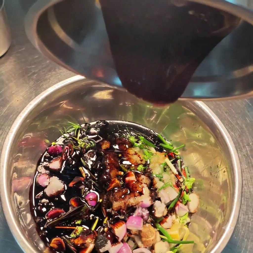 Salsa oscura siendo vertida desde un recipiente de acero en un bol lleno de varias hierbas, especias y aromáticos picados.