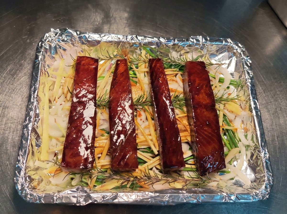 Cuatro bloques de atún marinados dispuestos ordenadamente en fila sobre una cama de jengibre triturado, cebollas, cebollinos y romero en una bandeja de horno forrada con papel de aluminio.