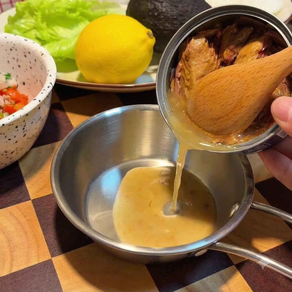 Una cuchara de madera retiene los trozos de atún dentro de una lata de metal mientras el aceite dorado se vierte en un pequeño bol de acero inoxidable.