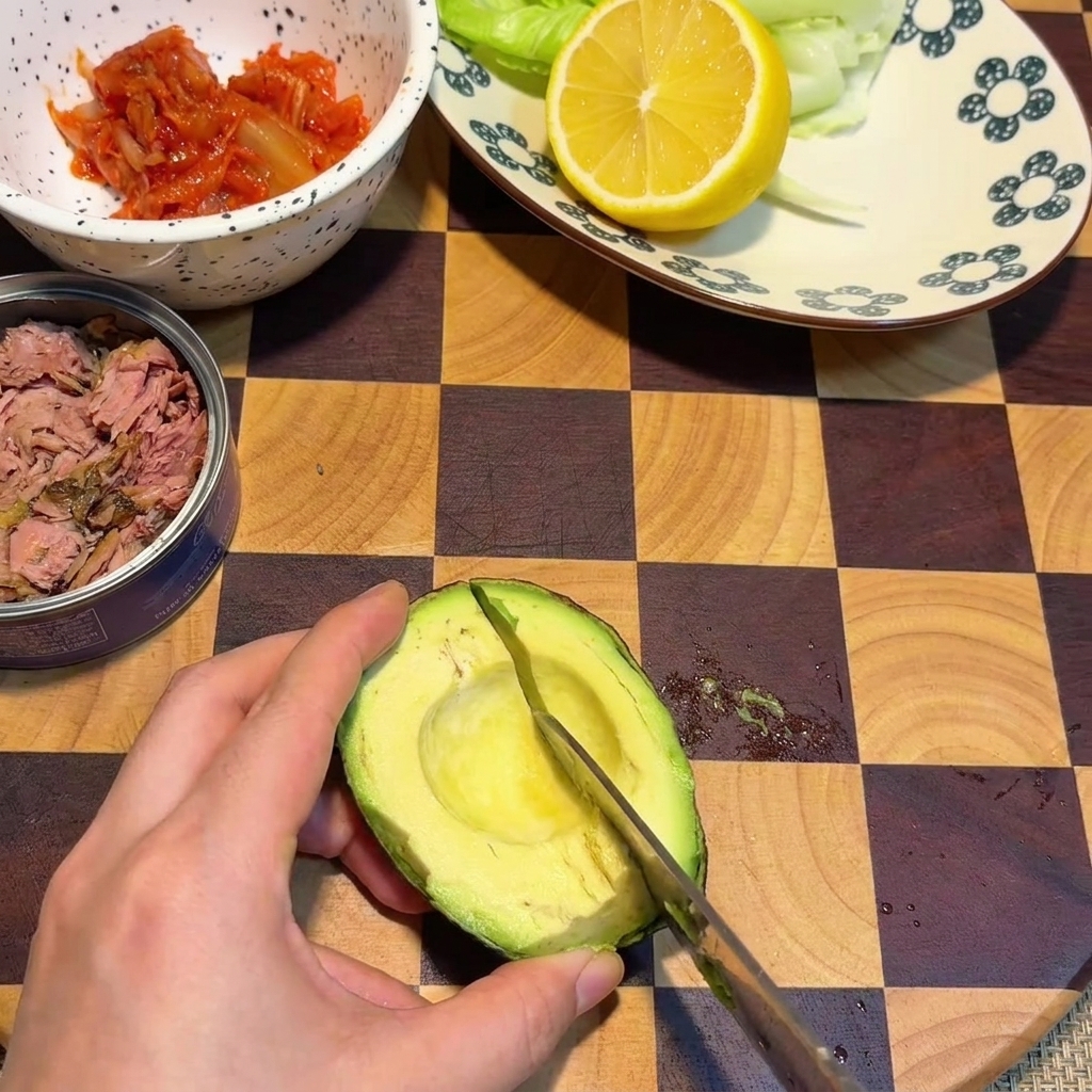 Una mano usando un cuchillo para cortar cubos en cuadrícula dentro de la mitad de un aguacate que descansa sobre una tabla de cortar de madera.