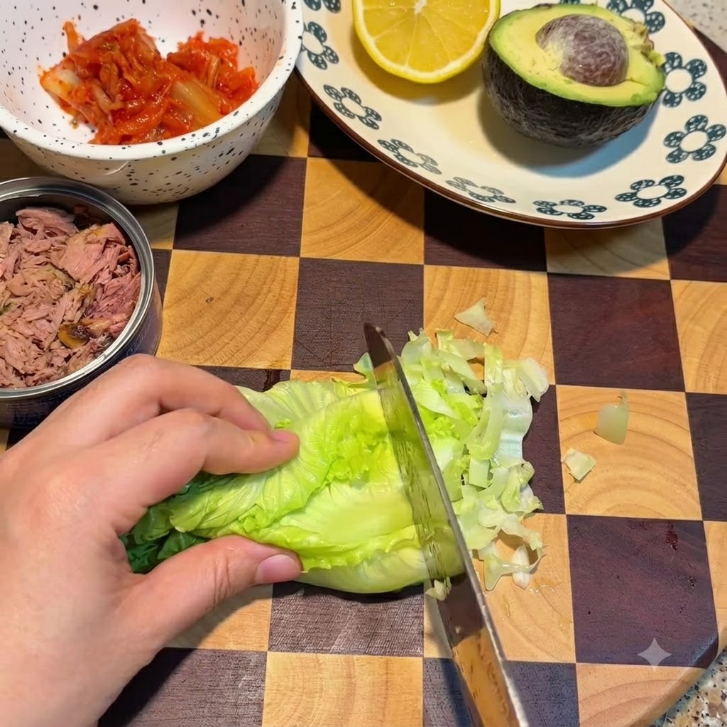 Un cuchillo cortando hojas de lechuga fresca verde en una tabla de cortar junto a un bol de kimchi y una lata de atún.