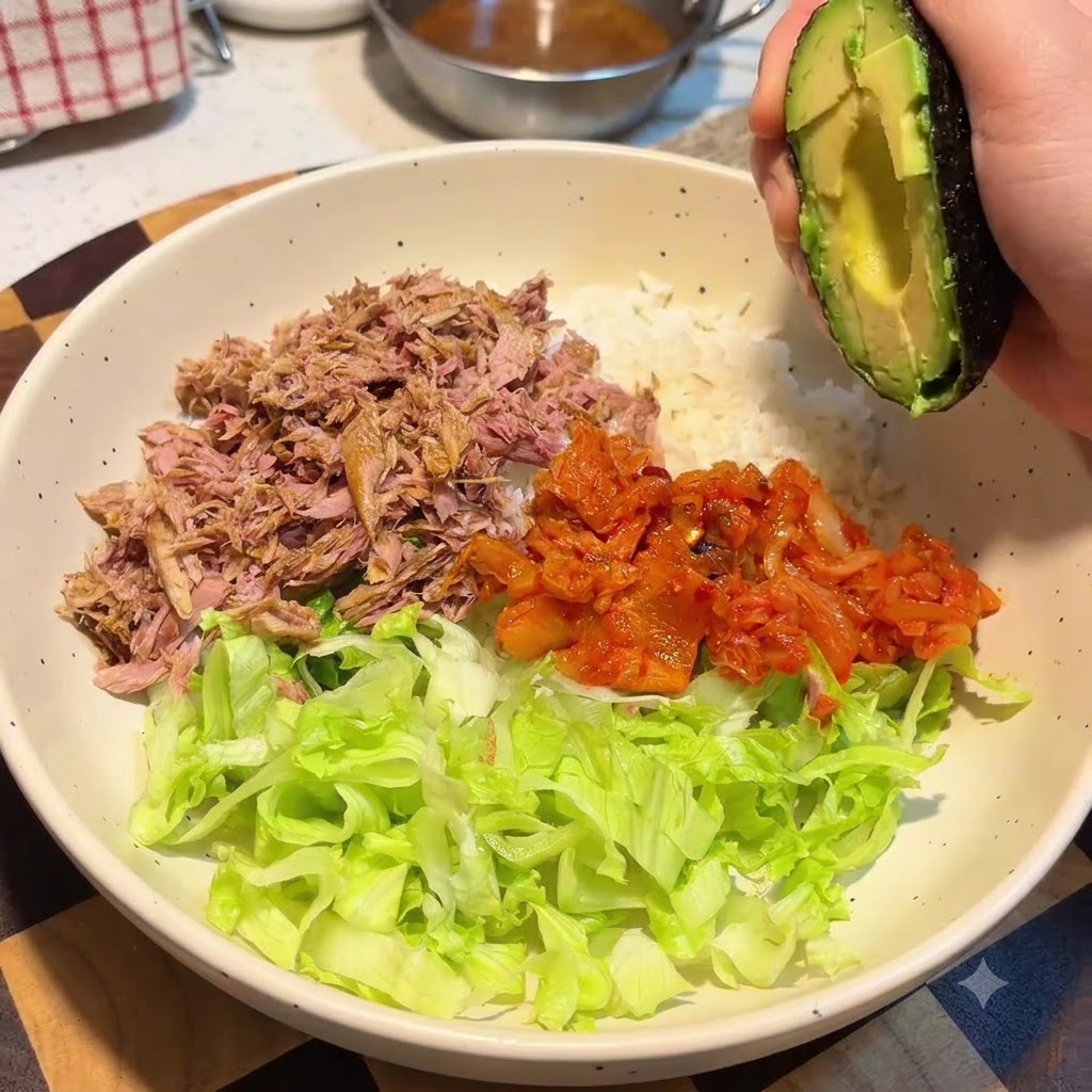 Una mano sosteniendo la mitad de un aguacate sobre un bol grande que contiene arroz, lechuga, atún y kimchi.