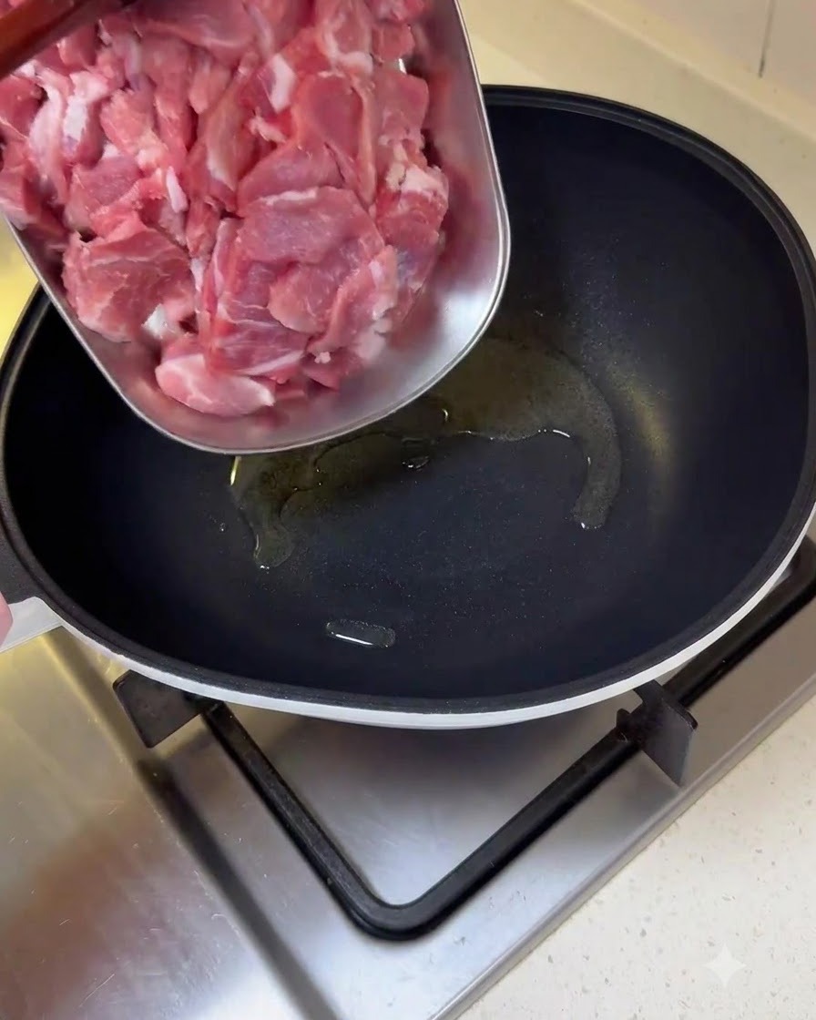 Trozos de cerdo crudo en rodajas siendo transferidos desde una bandeja a una sartén aceitada.