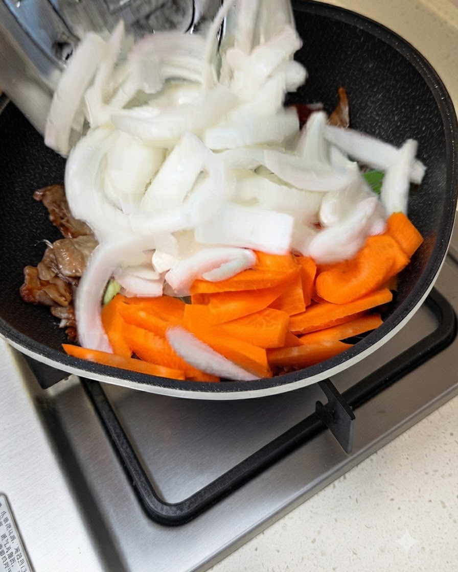 Cebollas blancas y zanahorias en rodajas siendo agregadas a una sartén con carne salteada y salsa.