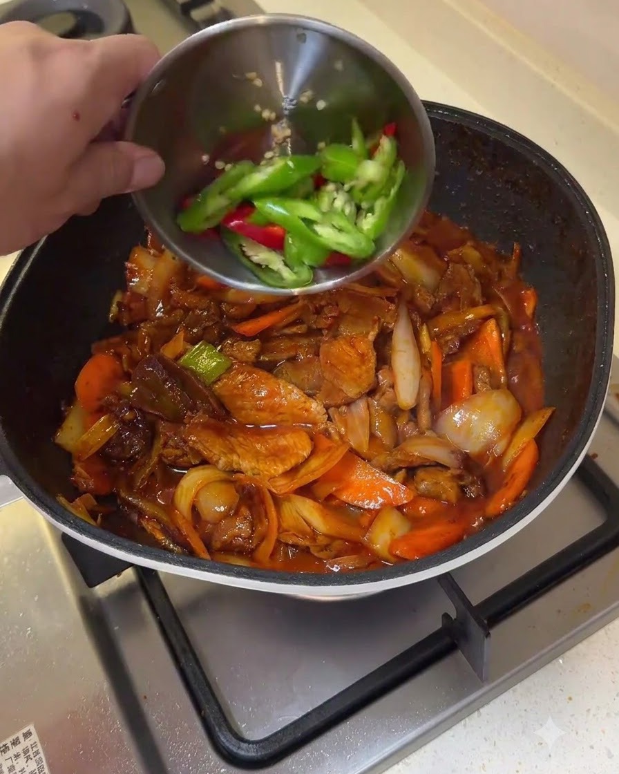 Rodajas de chile verde y rojo siendo vertidas desde un bol pequeño a una sartén de cerdo salteado y verduras.