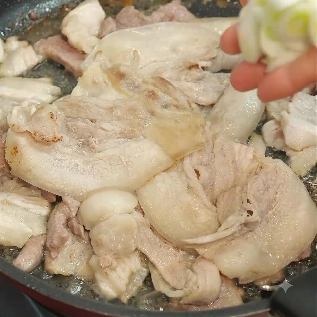 Una mano agregando cebollino picado en una sartén llena de rodajas de cerdo fritas.
