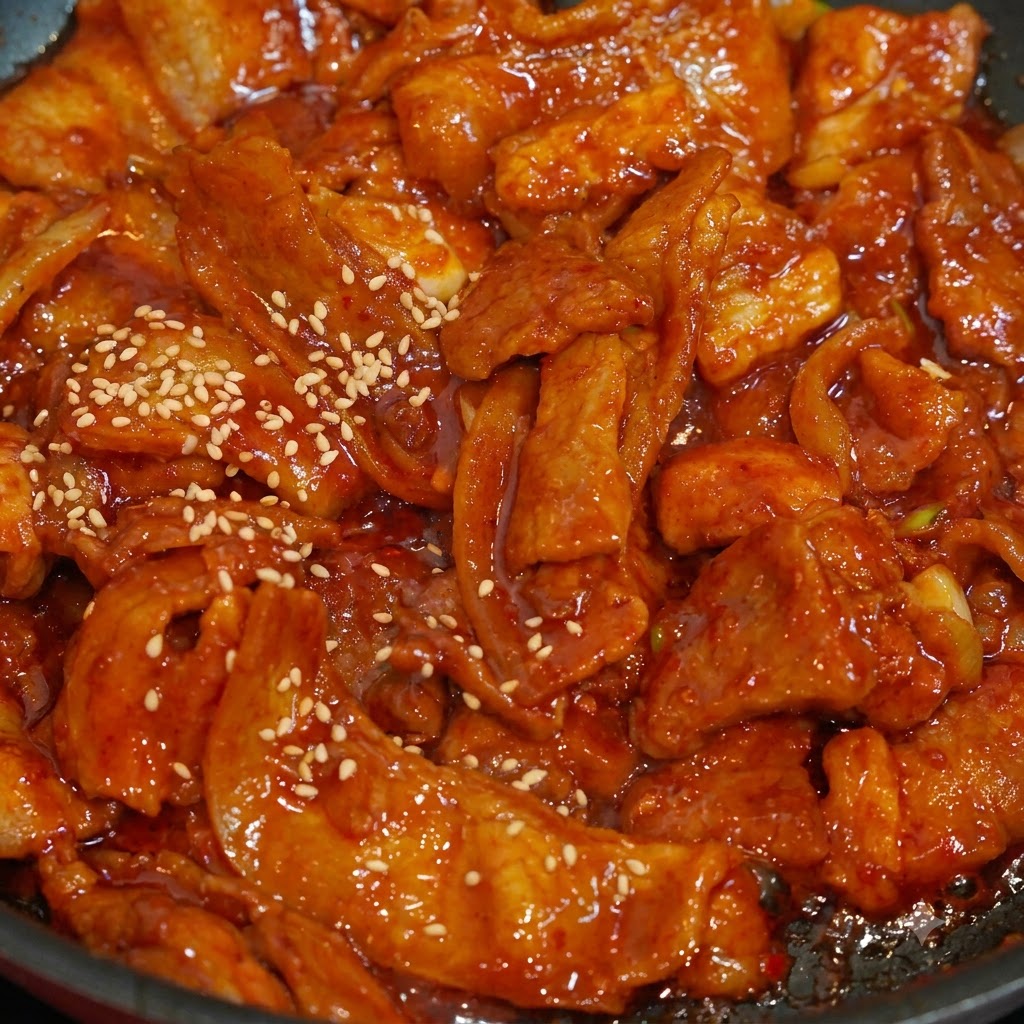 Una foto de cerca de semillas de sésamo blanco espolvoreadas sobre rodajas de cerdo picante con salsa roja en una sartén.