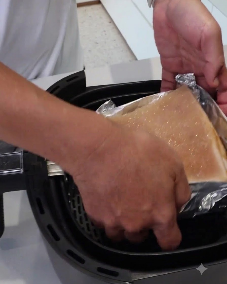 Un par de manos bajando un bloque de carne envuelto en papel de aluminio dentro de la cesta de la air fryer.