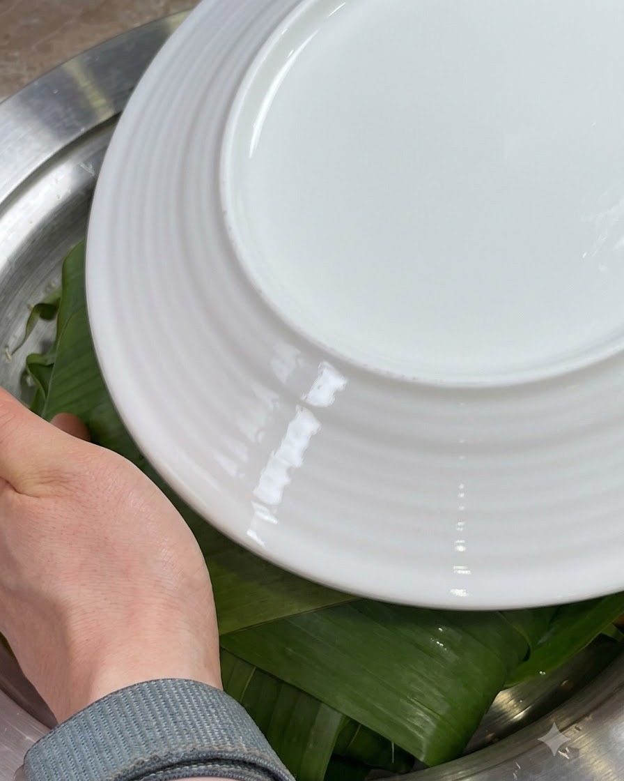 Manos doblando los bordes de las hojas de Zong verdes hacia adentro sobre los ingredientes, cubiertos con un plato blanco.