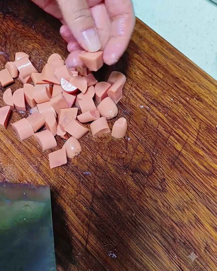 Manos cortando salchicha en trozos pequeños sobre una tabla de cortar de madera.