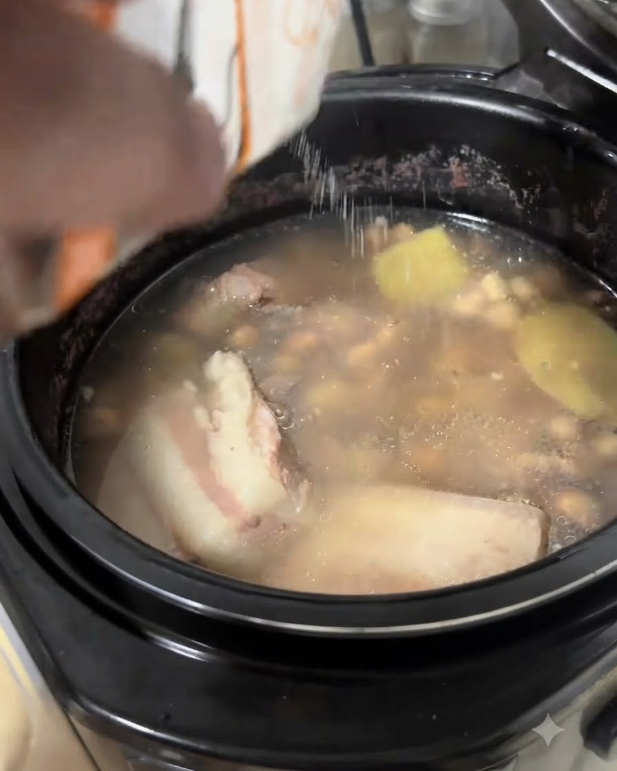 Sal siendo vertida desde un paquete blanco en una olla de sopa de costillas de cerdo y maní a fuego lento.