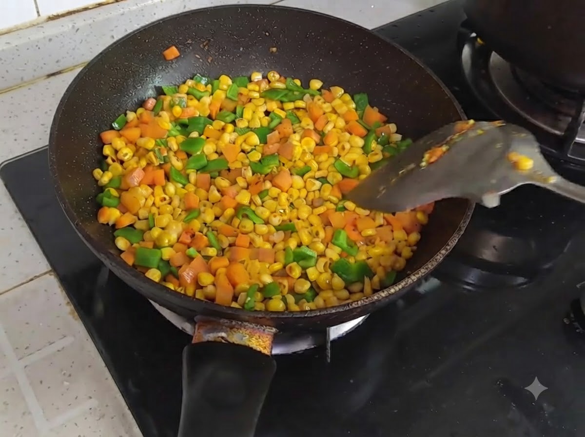 Una sartén llena de maíz, zanahorias y pimientos verdes cortados en daditos salteados y coloridos siendo removidos con una espátula.
