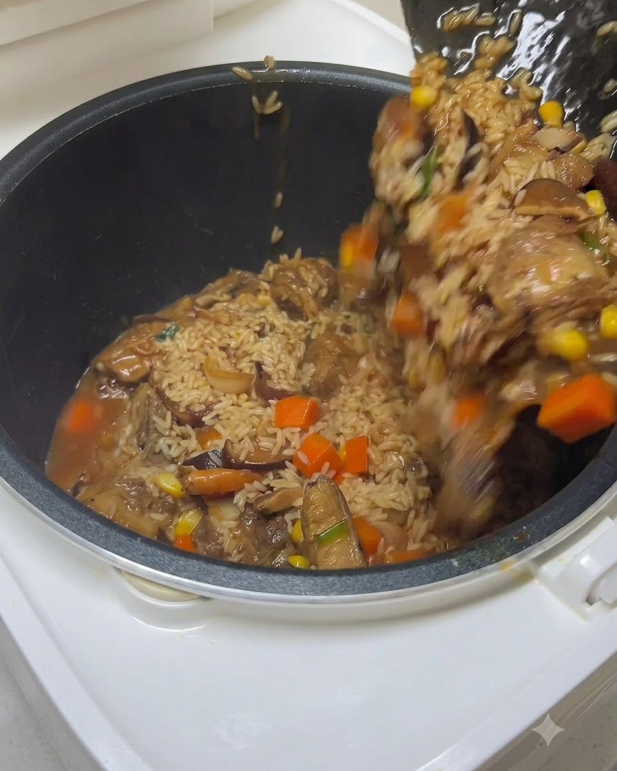 Una espátula transfiriendo arroz sazonado, costillas y verduras a la olla de la arrocera mientras se vierte agua.