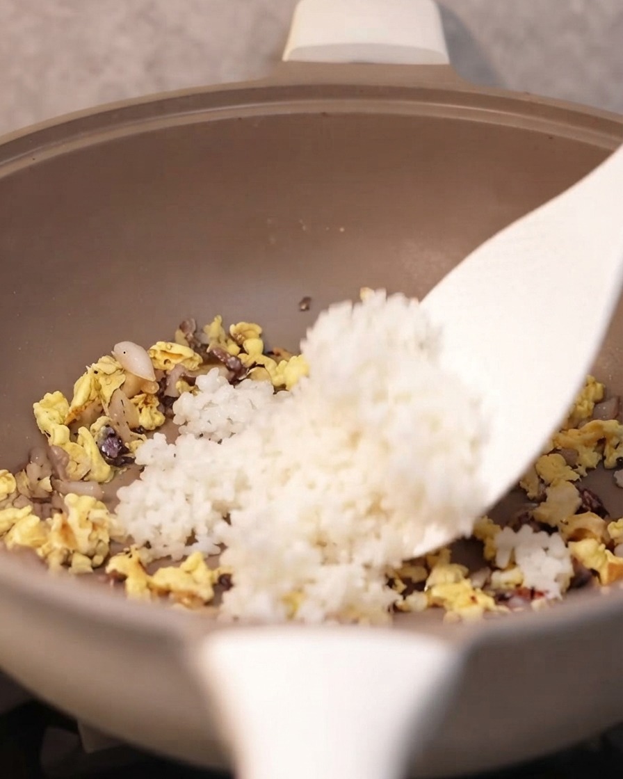 Una espátula añadiendo arroz blanco cocido en la sartén con los ingredientes sazonados.