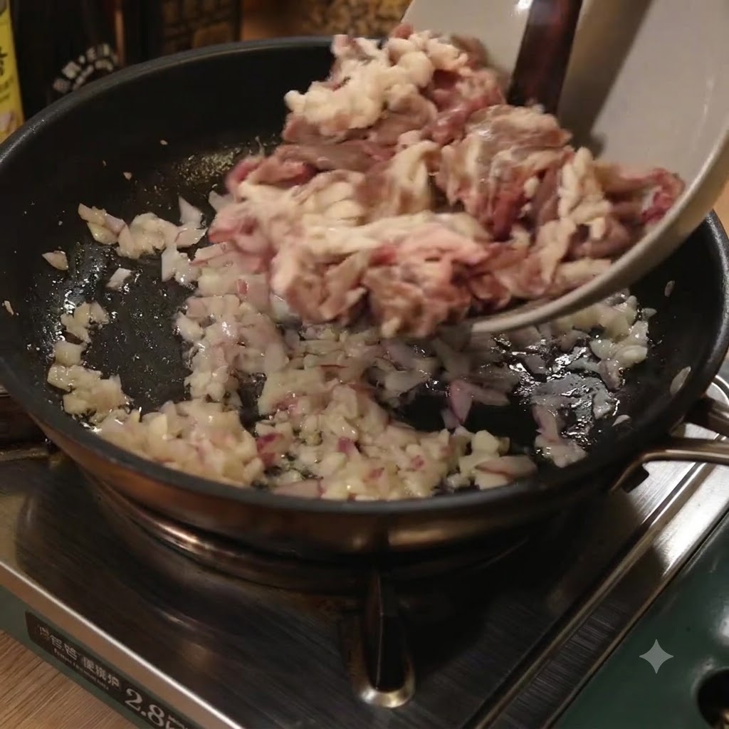 Tiras finas de carne agregándose desde un tazón a una sartén con cebollas y ajo salteados.