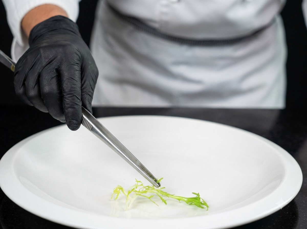 Un chef usando guantes negros y pinzas para colocar una guarnición de vegetales verdes frescos en el centro de un plato blanco grande.