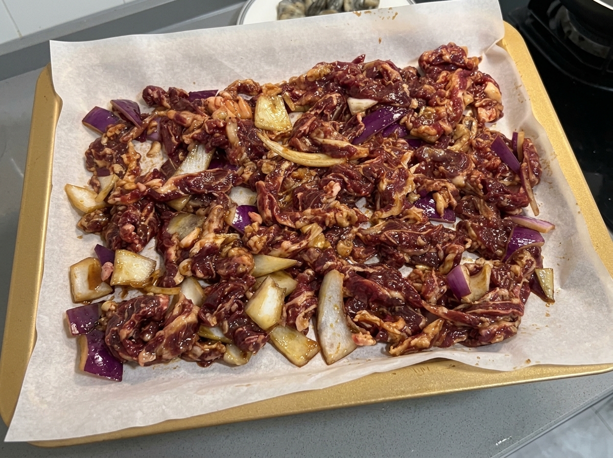 Ternera marinada y rodajas de cebolla distribuidas uniformemente en una bandeja de horno forrada con papel pergamino.