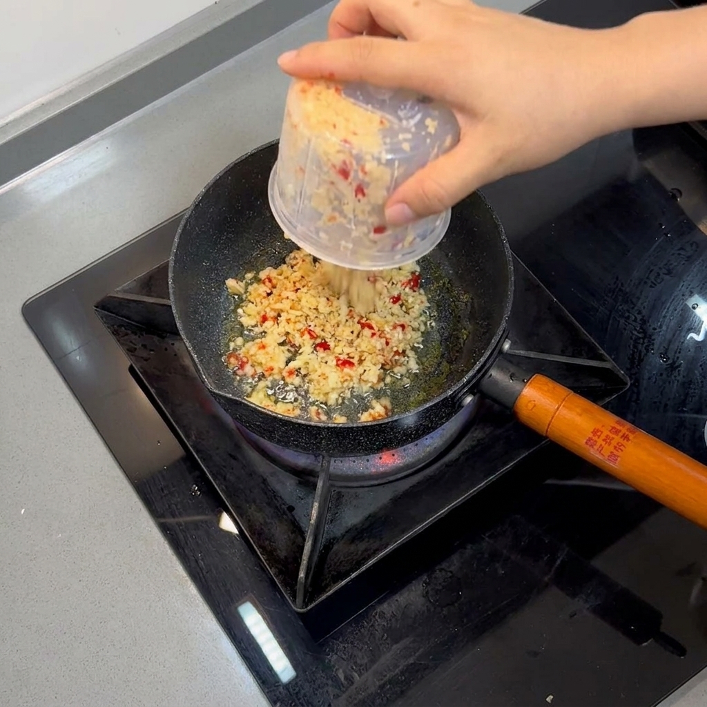 Ajo picado y chiles rojos friéndose en aceite caliente dentro de una sartén pequeña.