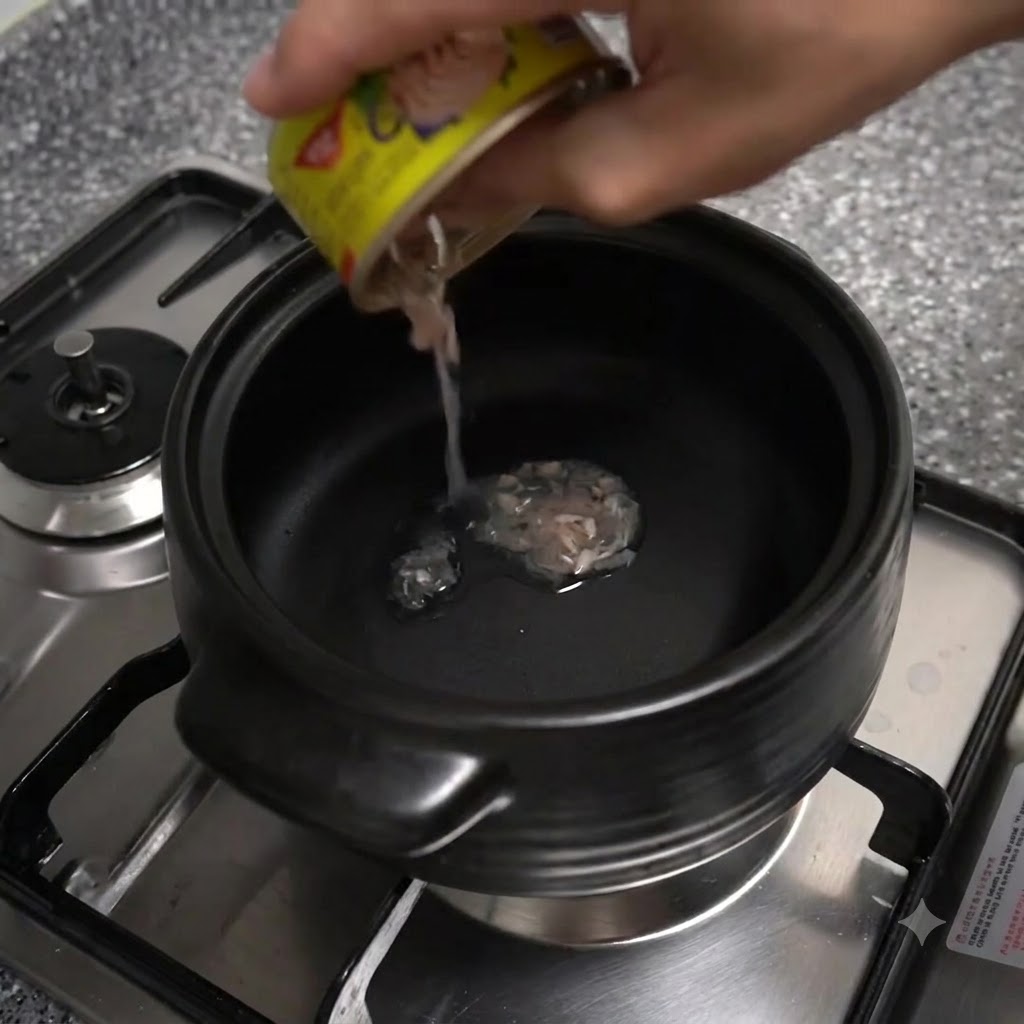 Aceite siendo vertido de una lata de atún pequeña con etiqueta amarilla en una olla de barro negra sobre un quemador de estufa.
