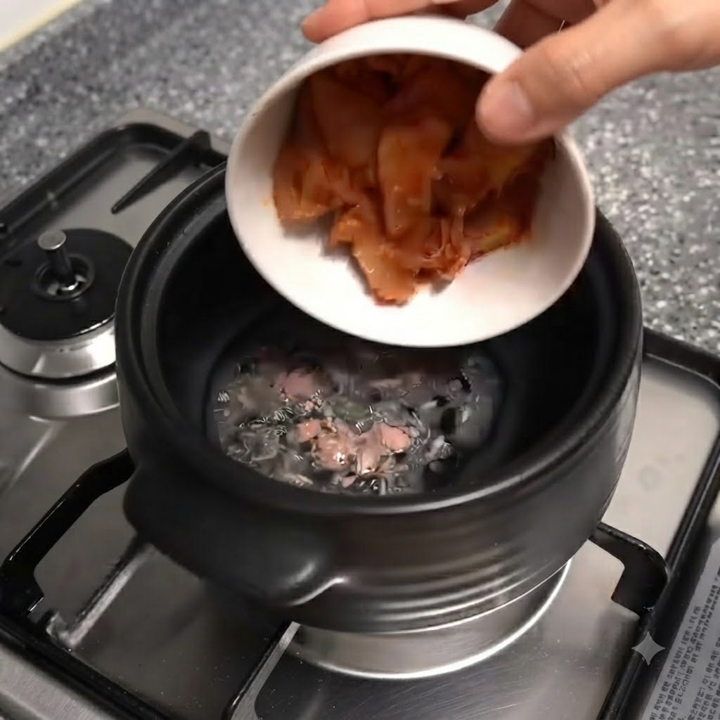 Un tazón blanco inclinándose y dejando caer trozos de kimchi rojo picado en la olla de barro negra.