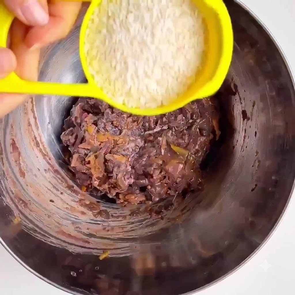 Harina de avena cayendo desde una taza medidora amarilla hacia un tazón de metal con frijoles machacados y verduras.
