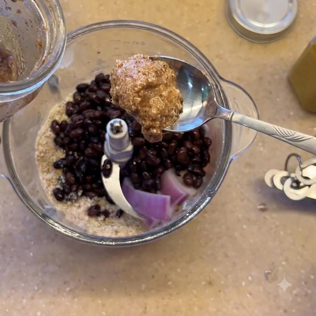 Una cucharada de mantequilla de almendras siendo sostenida sobre la licuadora que contiene frijoles negros y cebolla morada.