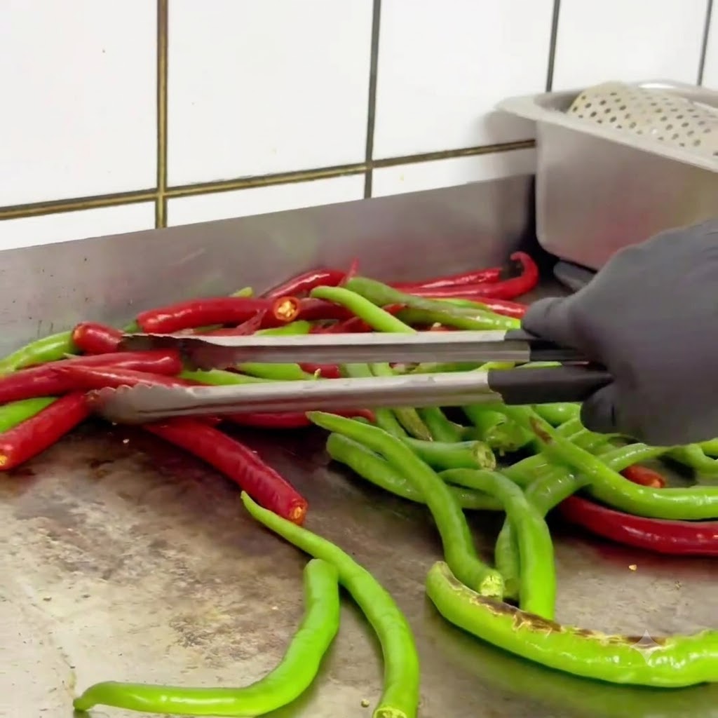 Chiles verdes y rojos siendo volteados con pinzas en una sartén caliente mientras se tuestan.