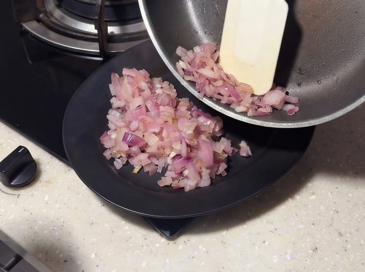 Cebollas salteadas deslizándose de una sartén a un plato negro usando una espátula.