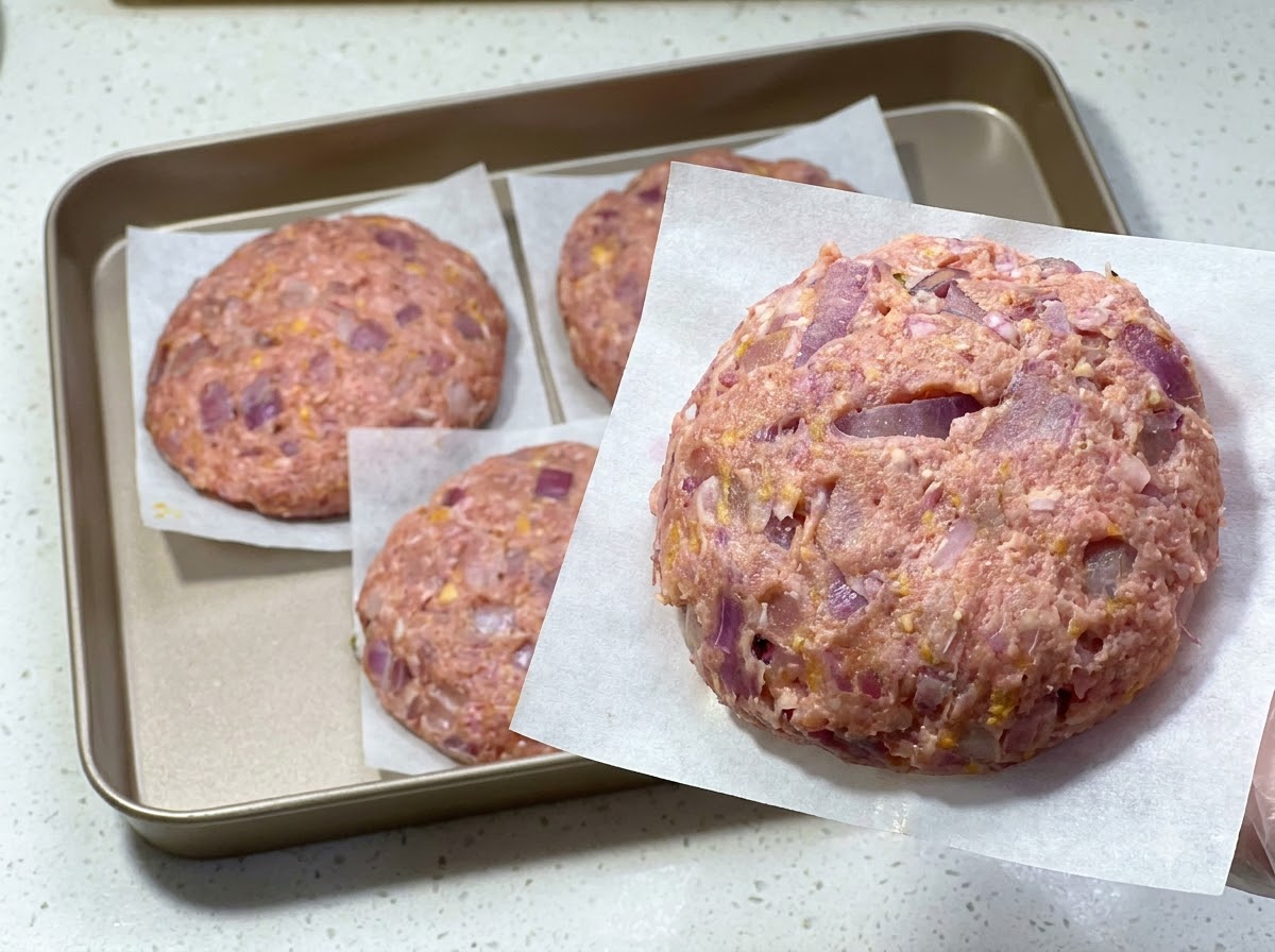 Una hamburguesa de carne cruda recién formada sobre papel pergamino, con otras hamburguesas colocadas en una bandeja detrás.