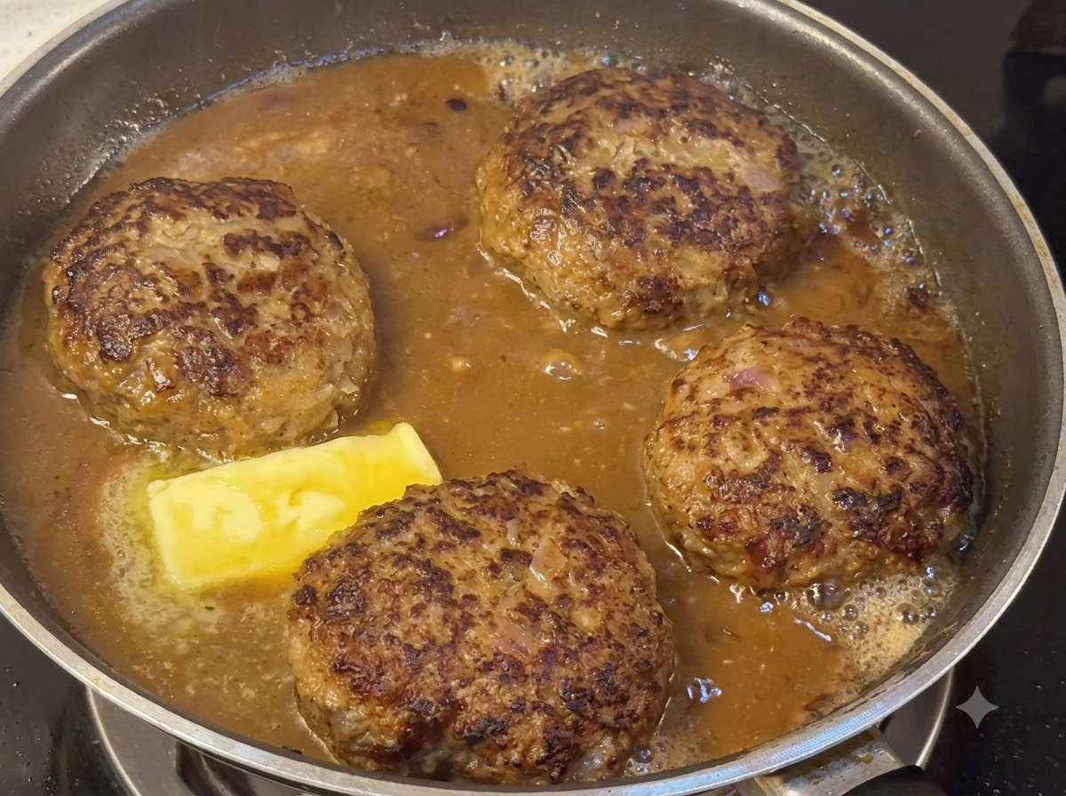Hamburguesas cociéndose a fuego lento en una espesa salsa marrón junto a un trozo de mantequilla derretida.