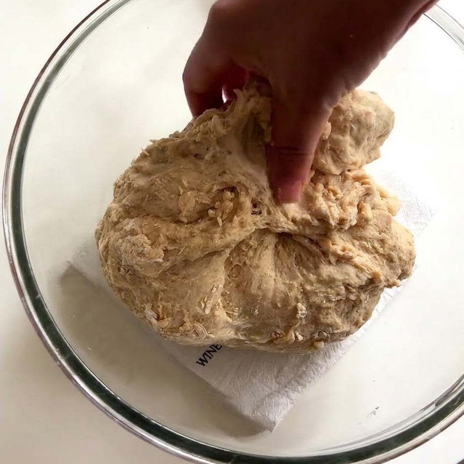 La mano de una persona usando dedos húmedos para estirar y jalar masa de pan pegajosa dentro de un tazón de vidrio.