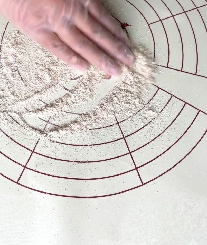 Una mano con guante esparciendo harina integral sobre una alfombrilla de silicona circular para amasar.