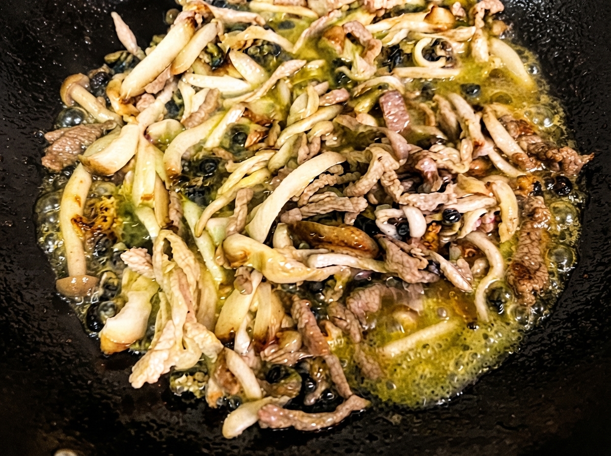 Setas shiitake picadas, setas enoki y caracoles de mar salteándose en aceite caliente dentro de un wok negro.