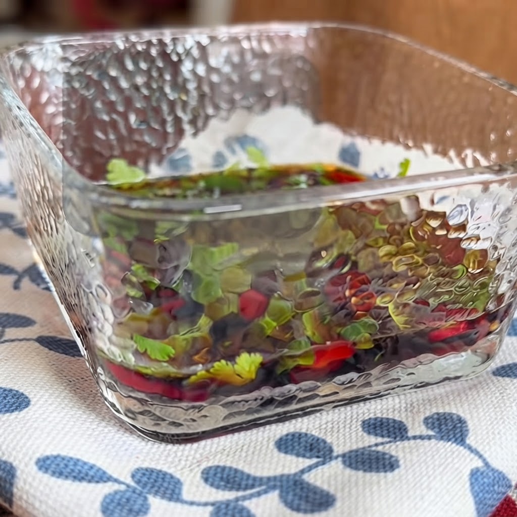 Un primer plano de un plato de vidrio cuadrado lleno de una salsa para untar salada que contiene hierbas y chiles picados mezclados con palillos.