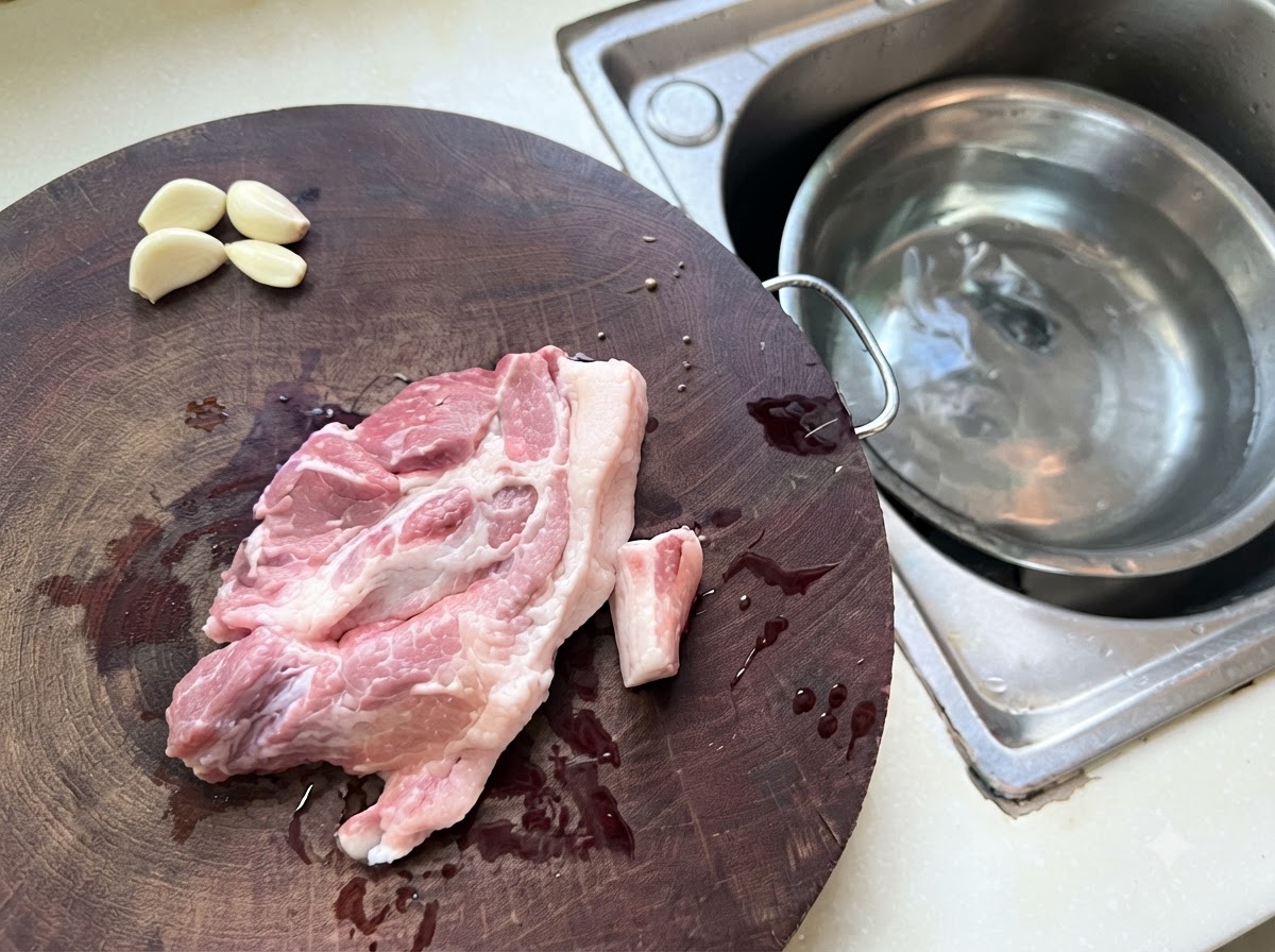 Trozos de cerdo picado y ajo distribuidos en una tabla de cortar de madera al lado de un tazón de metal con agua.