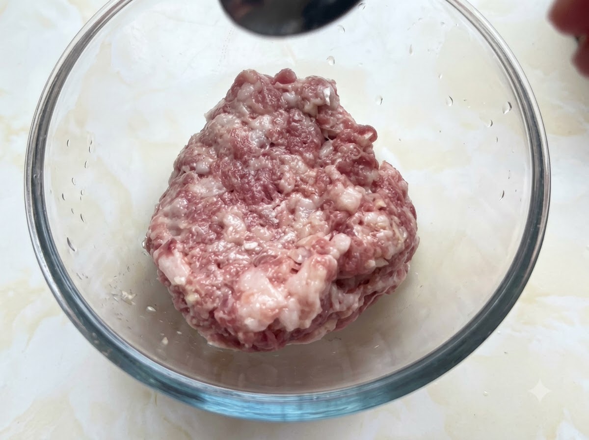 Un tazón de vidrio lleno de cerdo picado recibiendo condimentos de una cuchara sostenida arriba.