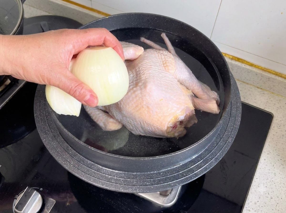 Mitad de cebolla y otros aromáticos siendo arrojados a una olla que contiene un pollo entero y agua.